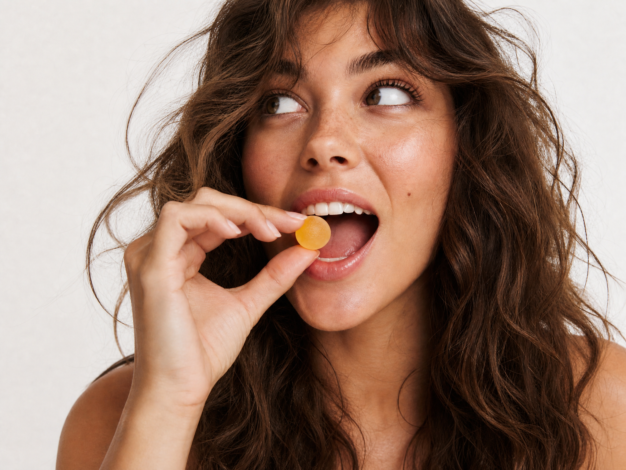 A close-up of a woman with long wavy brown hair holding a small round yellow gummy candy near her open mouth, set against a plain light background.