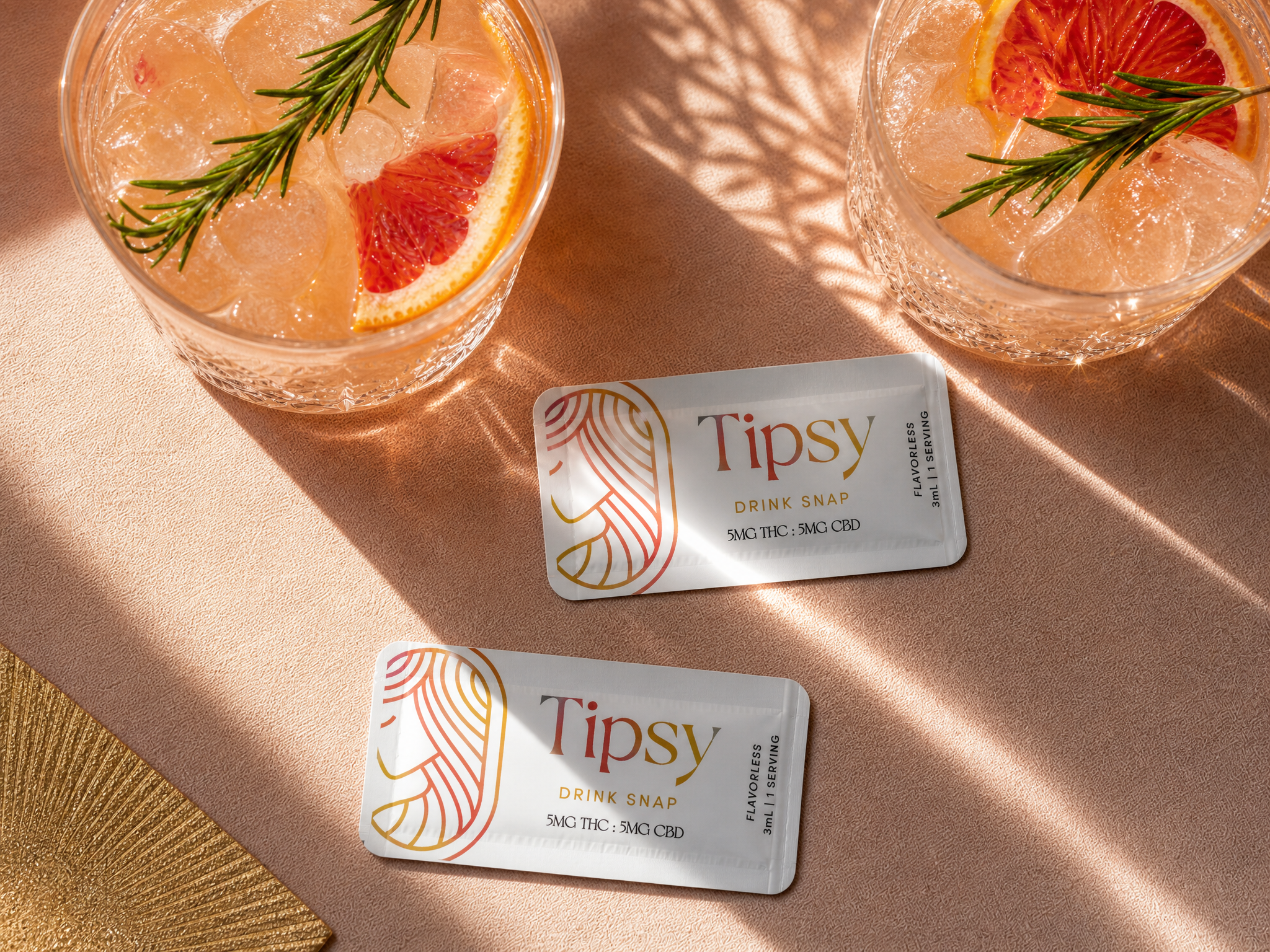 Two Tipsy Drink Snap cannabis tincture packets are arranged on a beige surface between two glasses of pink citrus cocktail with rosemary and grapefruit garnish, under warm dappled sunlight.